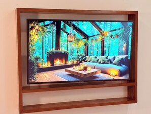Un televisor en marco muestra una habitación acogedora con chimenea, sofá y plantas en macetas.