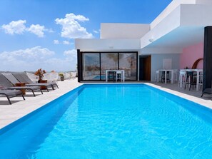 Un espace piscine moderne avec une piscine à l’eau d’un bleu limpide, des chaises longues et un coin salon équipé de tables et de chaises.