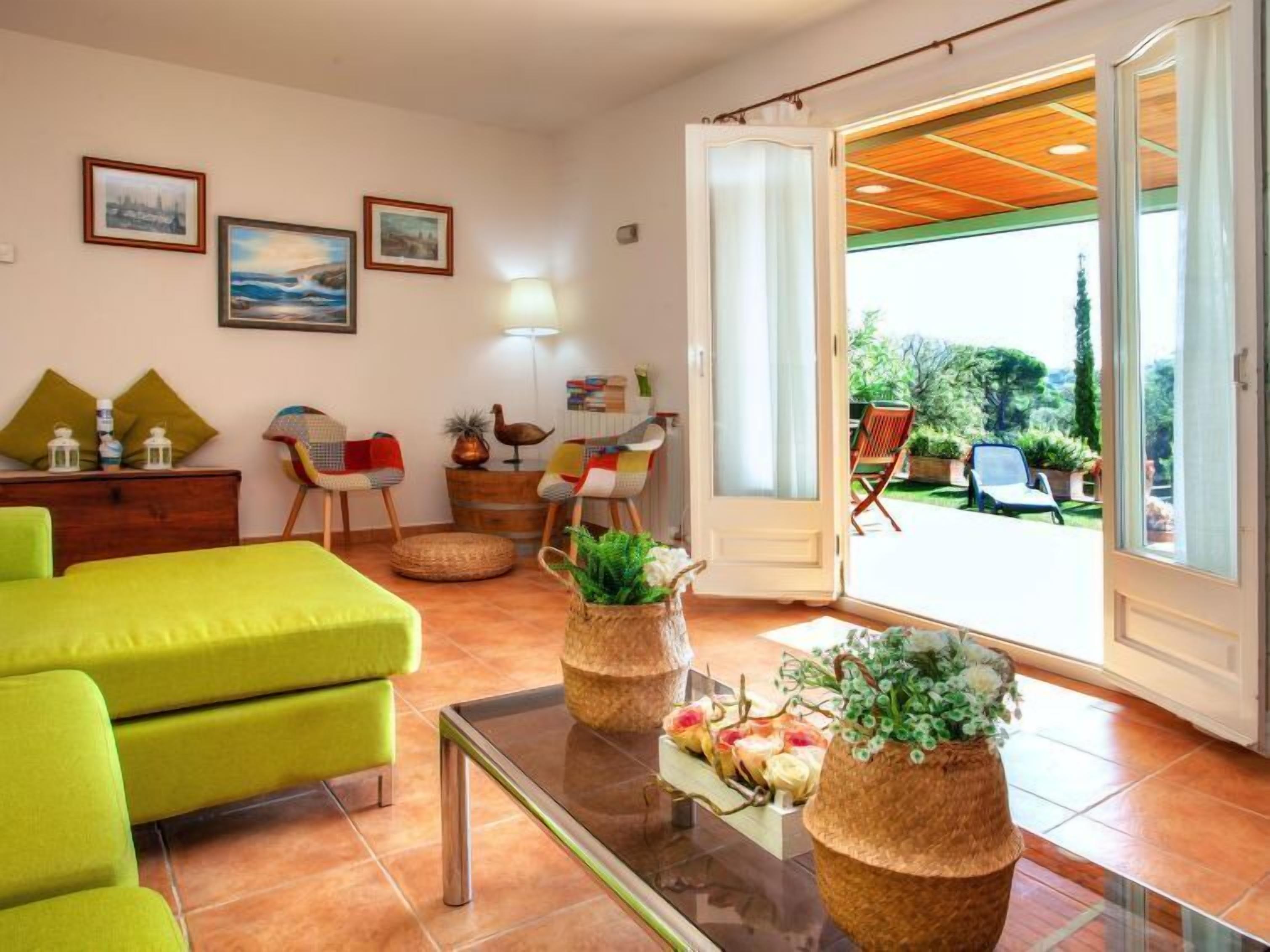 A cozy living room with a green sofa, a glass coffee table, and wicker vases with flowers. There are two chairs, a lamp, and a door leading to a balcony with outdoor furniture.