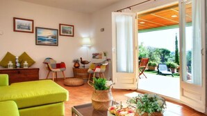 Ein gemütliches Wohnzimmer mit einem grünen Sofa, einem Glastisch und Korbsvasen mit Blumen. Zwei Sessel, eine Lampe und eine Tür zu einem Balkon mit Gartenmöbeln sind ebenfalls vorhanden.