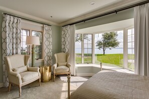 A bedroom with a large bed, two armchairs, a side table, and a view of a lake. - Azaleana Manor (Orange Park)