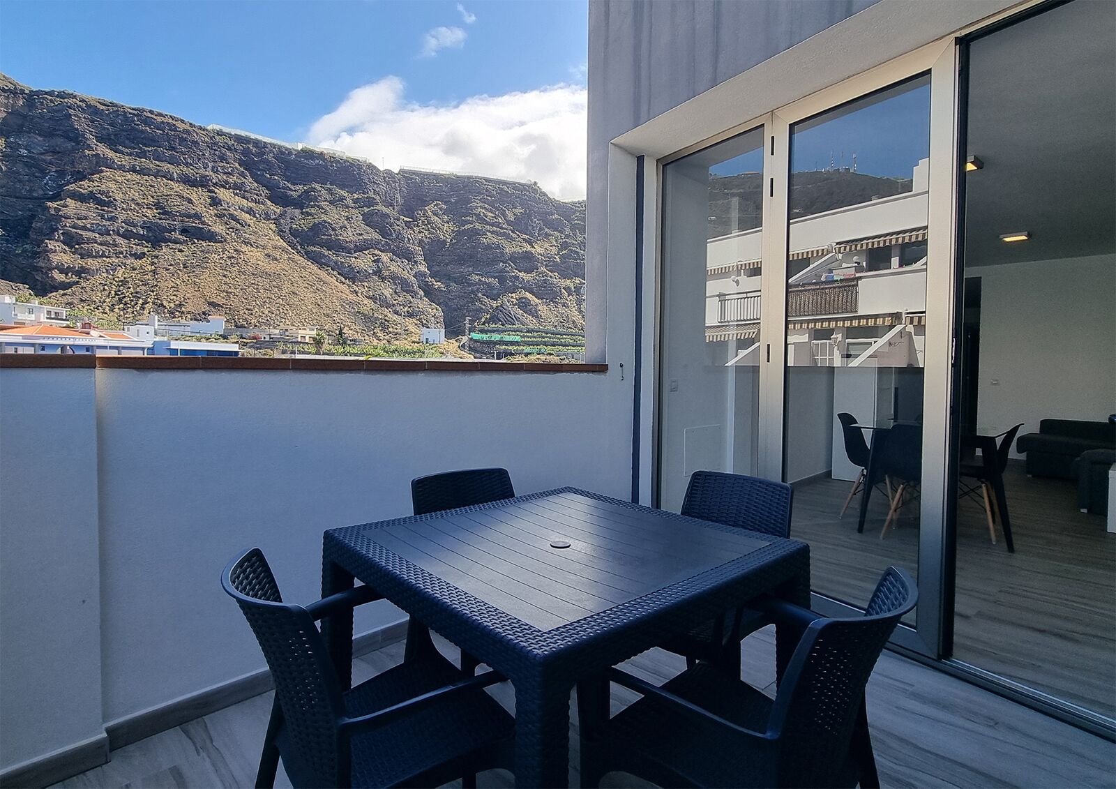 A balcony with a table and chairs, a sliding glass door leading to an indoor area, and a view of mountains and buildings in the background.