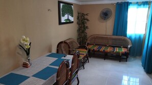 Un comedor con una mesa preparada para cuatro personas, rodeada de sillas de madera y un banco. Un ventilador está montado en la pared, y se ve una planta en maceta.