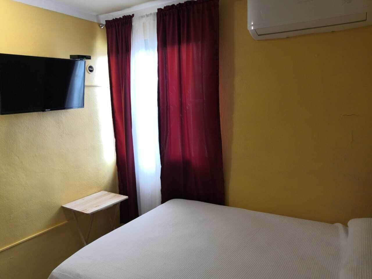 A hotel room with a bed, a small bedside table, a TV mounted on the wall, and a window with red curtains.