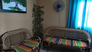 Una habitación con dos sofás de mimbre con cojines de colores, un ventilador de pie, una planta de bambú y un cuadro de un árbol en la pared.