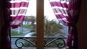 Una ventana con cortinas a rayas moradas y blancas, una barandilla metálica y una vista de una carretera con palmeras y personas a lo lejos.