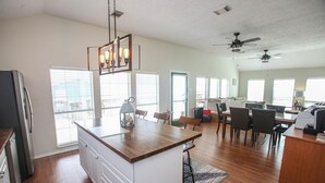 A spacious kitchen with a wooden island, white cabinetry, and a dining area with a table and chairs. There are ceiling fans, a refrigerator, and a window with blinds.