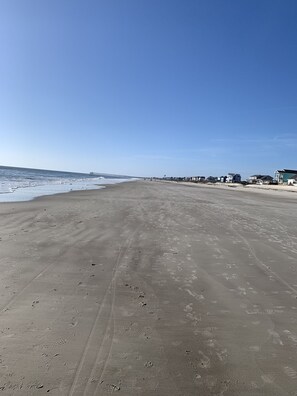 Uma extensa praia de areia com pegadas e marcas de pneus, céu azul límpido e edifícios ao fundo.