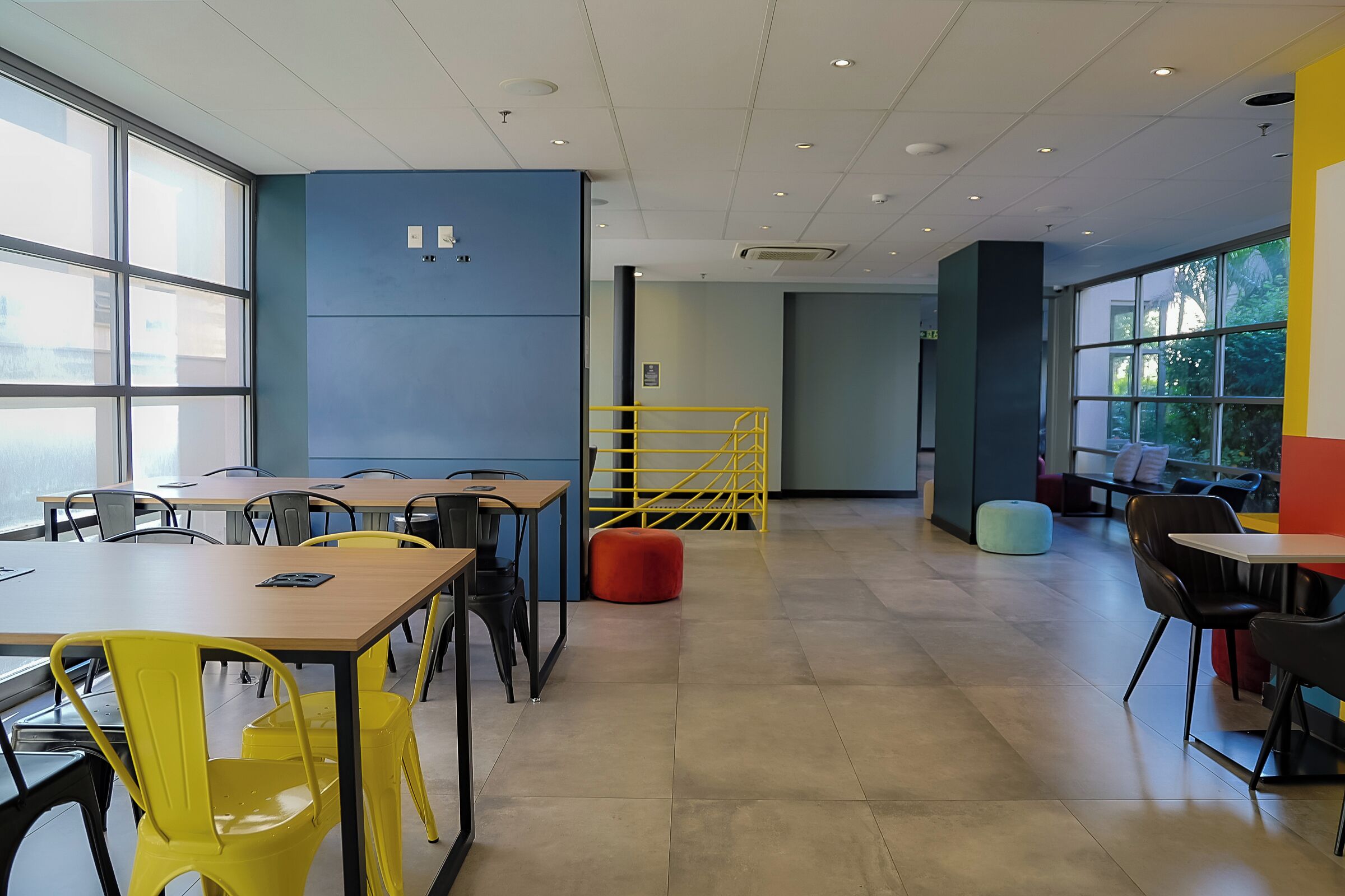Un espacio interior moderno con mesas, sillas, una escalera y ventanas amplias.