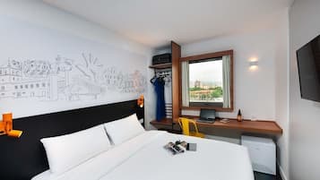 A hotel room with a bed, a desk with a computer, a chair, a TV, and a window with a view of buildings.