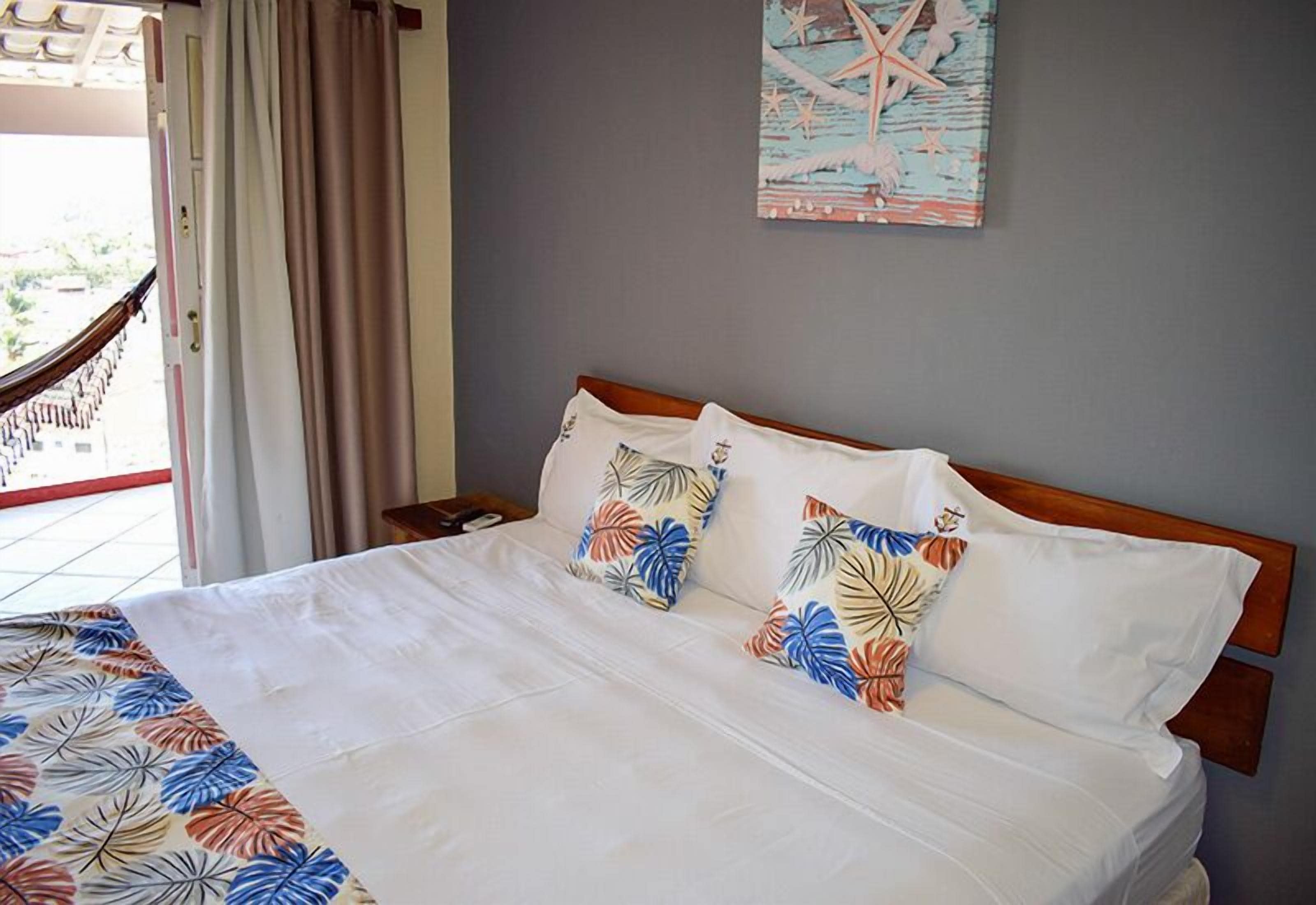 A hotel room with a bed, a nightstand, a window with curtains, and a wall with a nautical-themed painting.