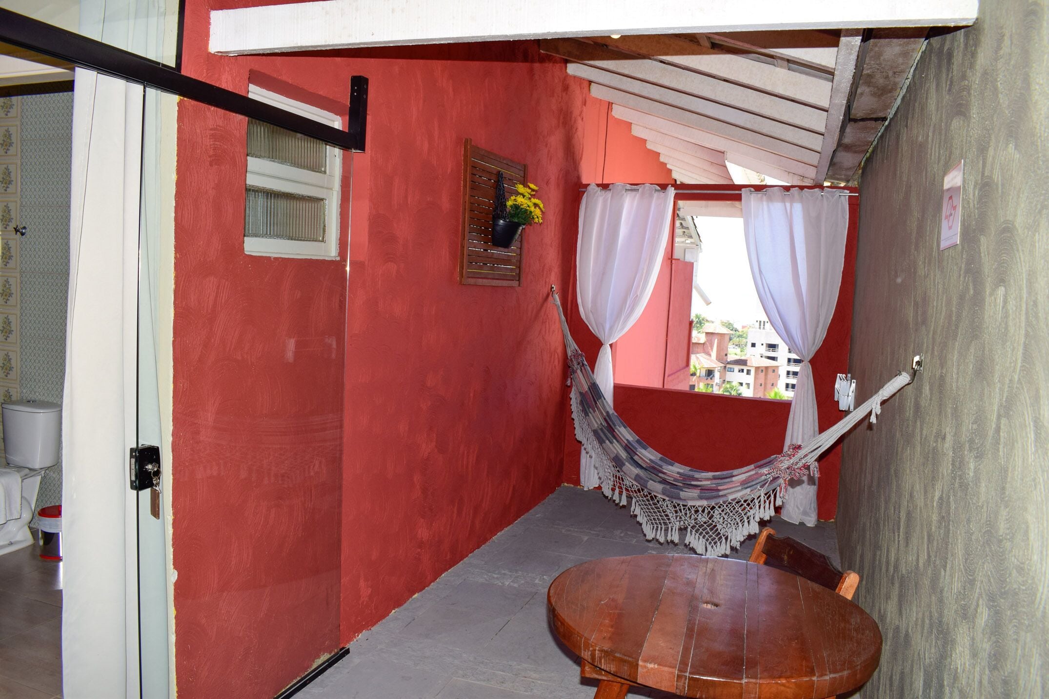 A red-walled room with a hammock, a wooden table, and a window with a curtain.