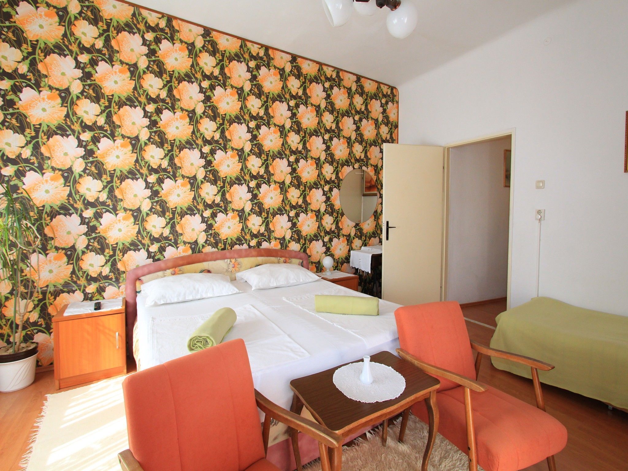 A hotel room with a floral wallpaper, a bed with white linens, two orange chairs, a small table with a vase, and a green sofa.