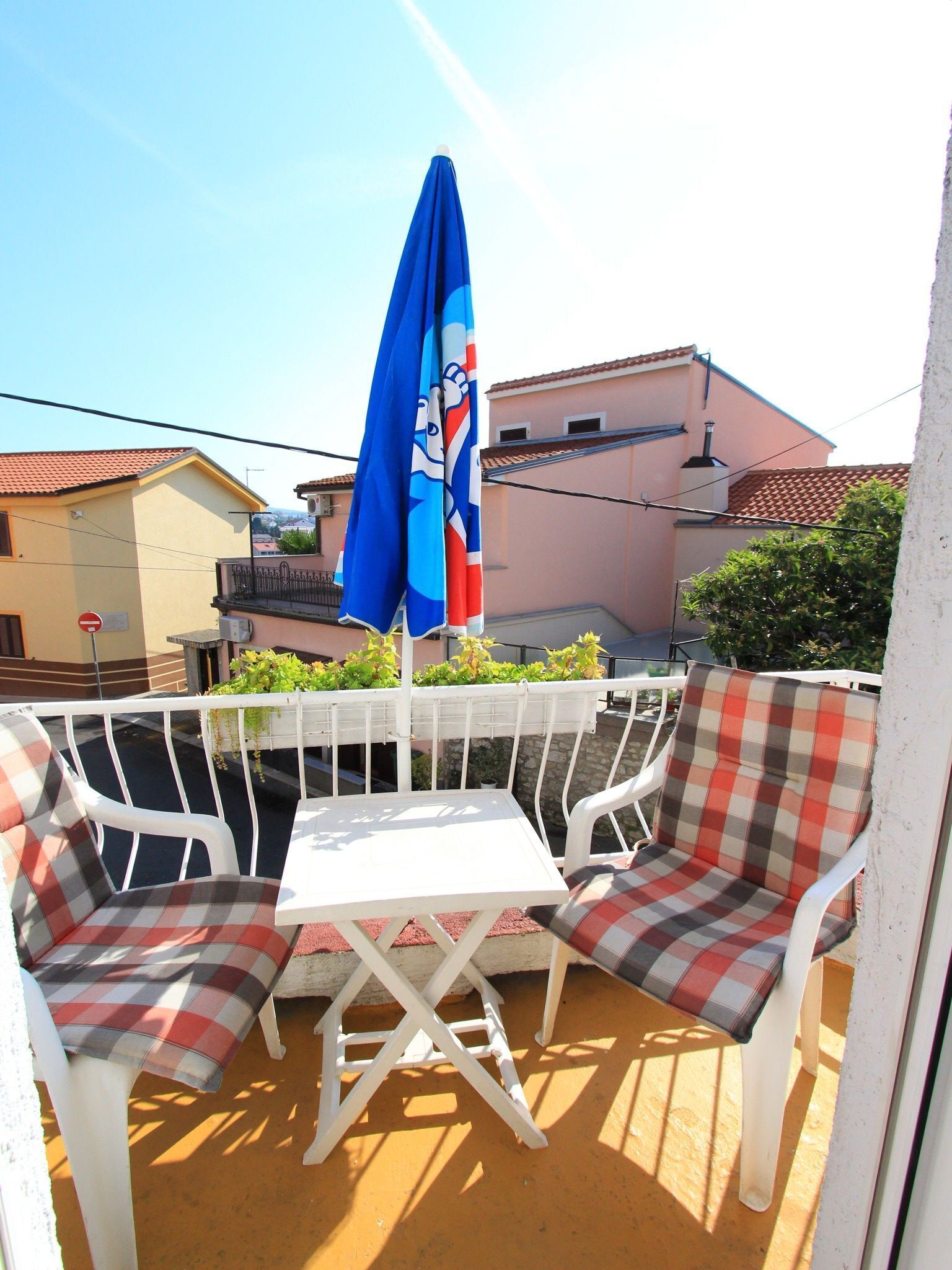 A small outdoor seating area with a table and two chairs, a blue umbrella, and a view of buildings in the background.