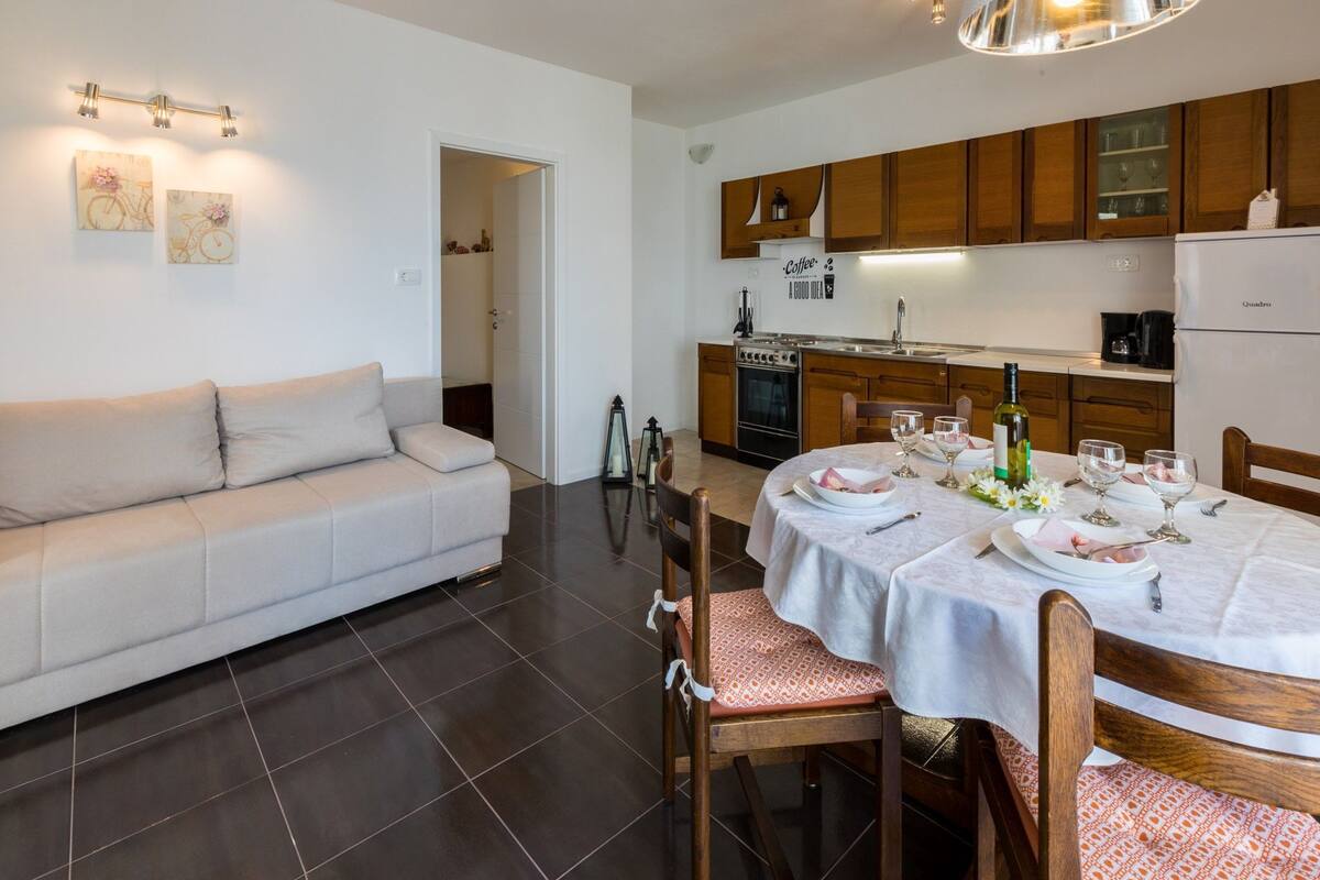 Una cocina moderna con armarios de madera, horno empotrado y refrigerador. Hay una mesa de comedor con platos, vasos y una botella de vino. Un sofá blanco está apoyado contra la pared, y hay luces decorativas en la pared sobre la encimera de la cocina.