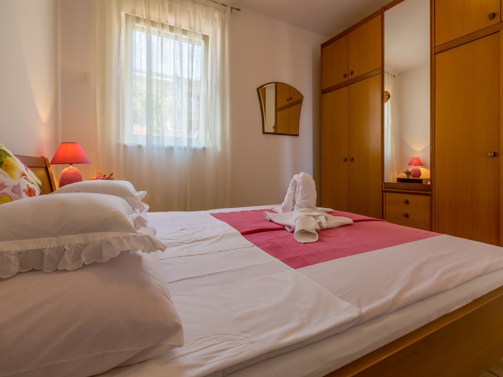 Una cama bien hecha con sábanas blancas, un edredón rosa y una toalla sobre un tapete rosa. Hay un armario de madera con espejo, una mesita de noche con lámpara y una ventana con cortinas traslúcidas.
