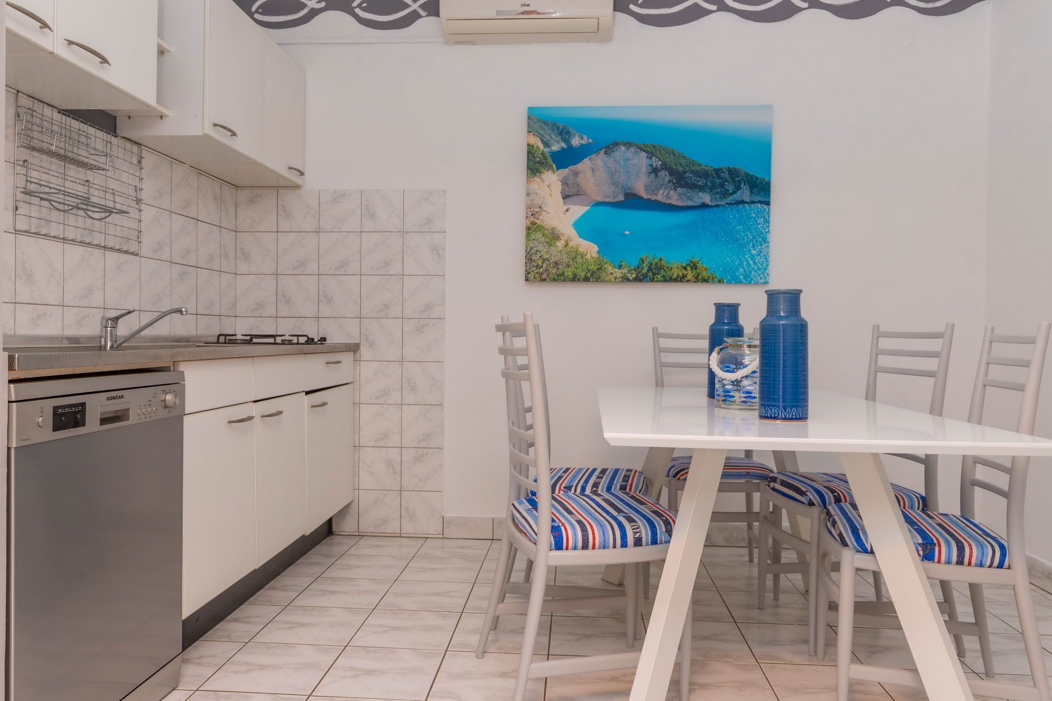 Cozinha com armários brancos, lava-louças e uma pintura de paisagem costeira.