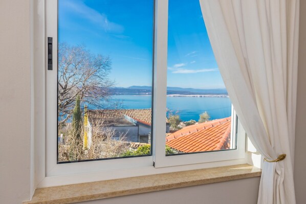 A window with a view of a coastal landscape, including a body of water and buildings with red-tiled roofs.