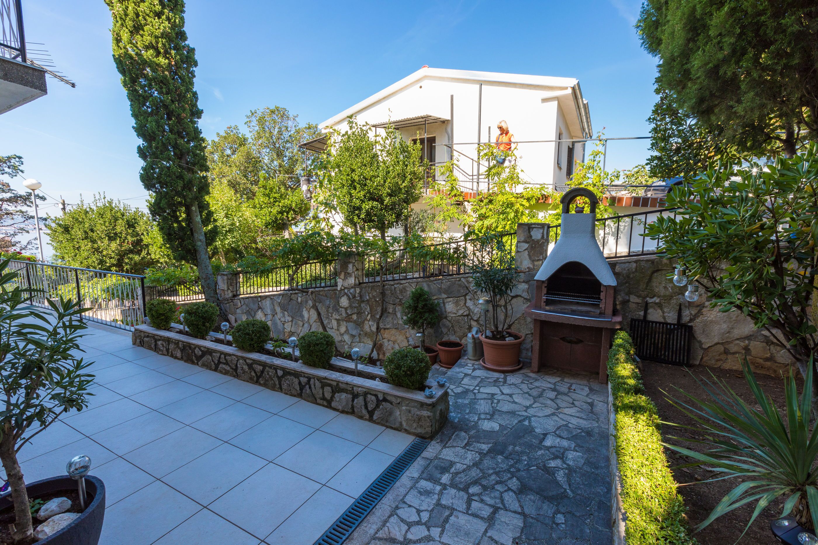 A courtyard with a stone pathway, potted plants, a stone wall, and a building with a balcony.