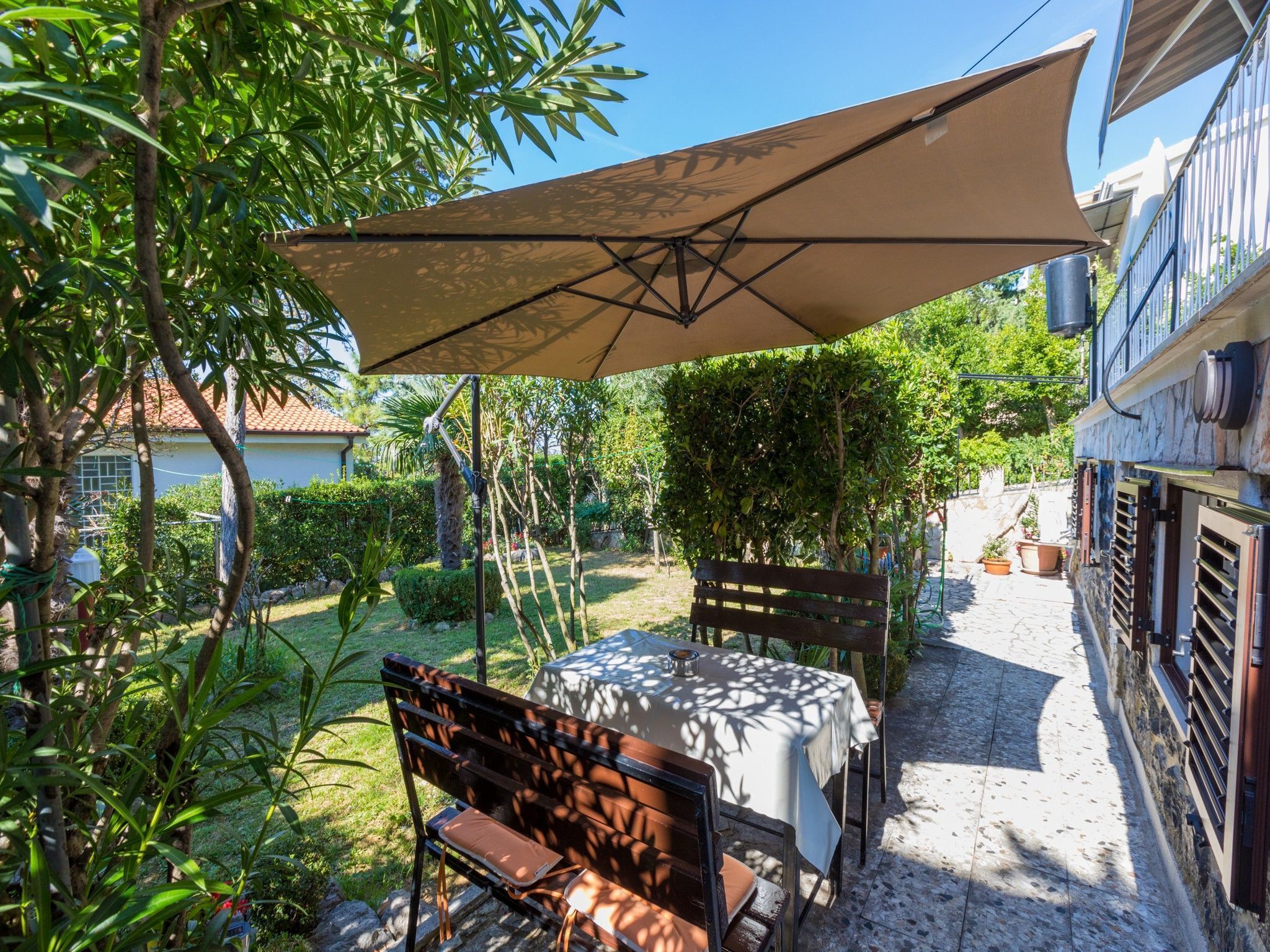 En udendørs terrasse med et bord og stole under en stor parasol, omgivet af grønne planter og en stentrappe.