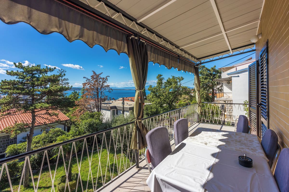 A balcony with a table and chairs, overlooking a scenic view of the sea and surrounding buildings.