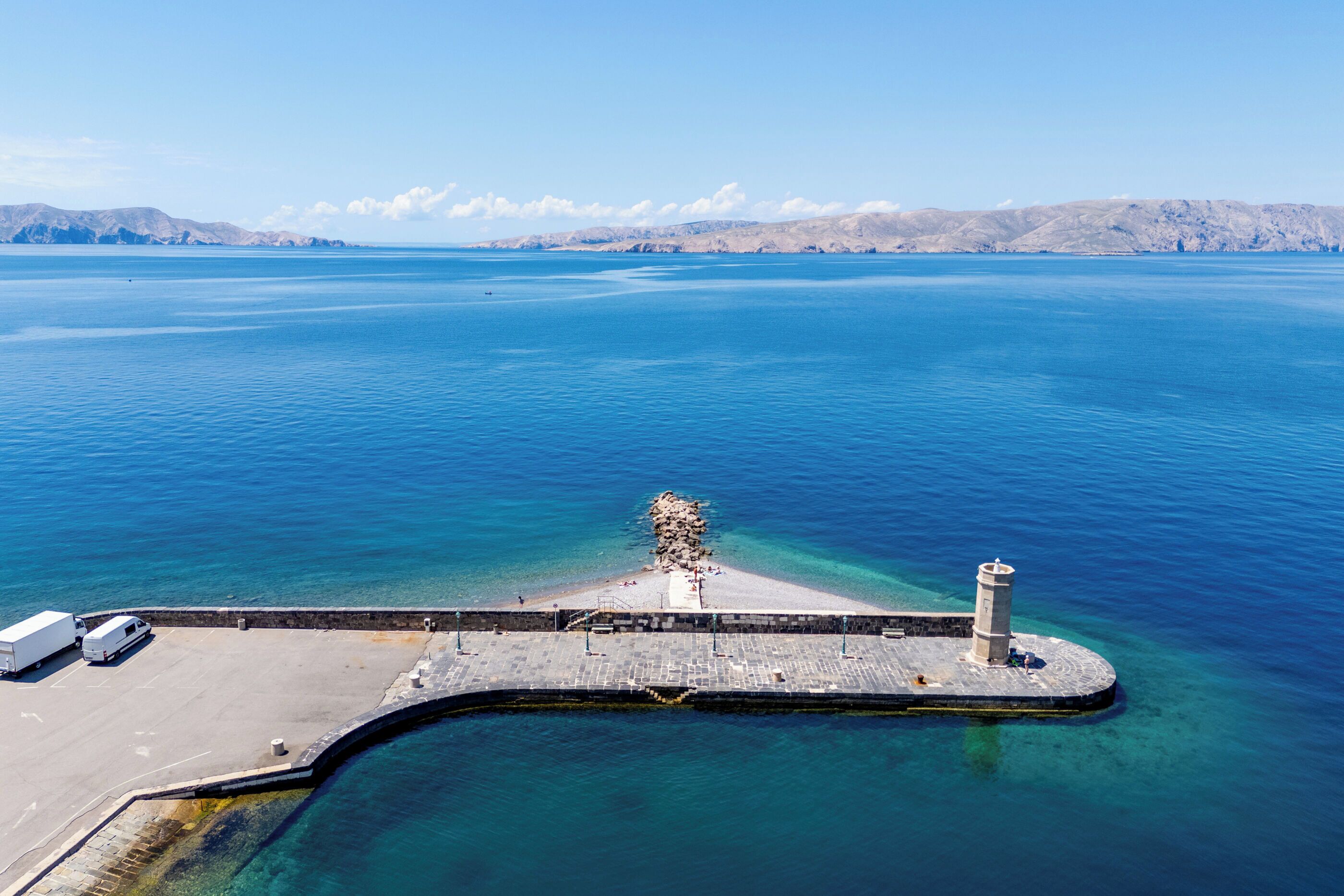 Vista aérea de una zona costera con un muelle que se adentra en el agua, un faro y un estacionamiento con vehículos.