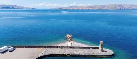 Vista aérea de una zona costera con un muelle que se adentra en el agua, un faro y un estacionamiento con vehículos.