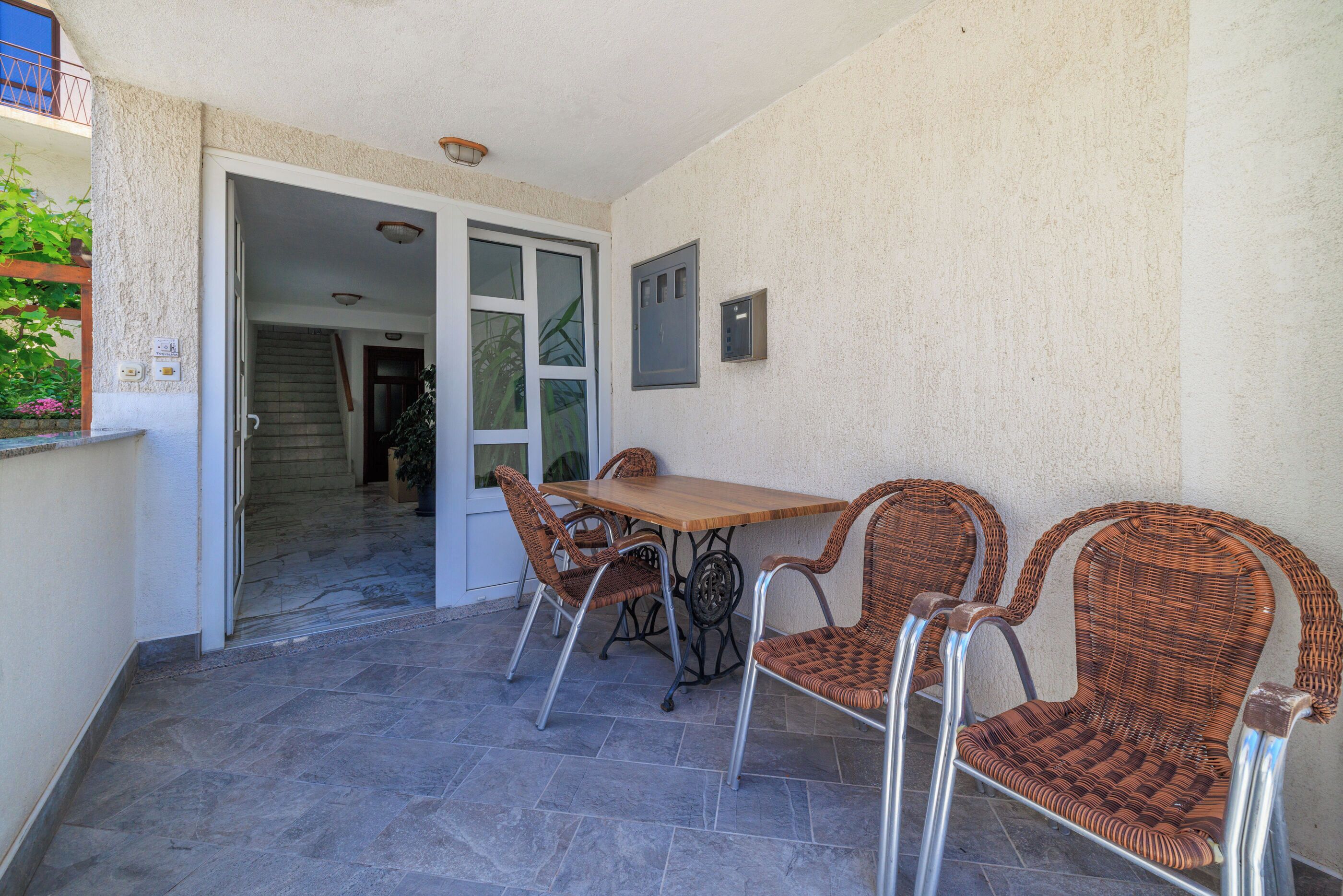 A patio area with a wooden table and wicker chairs, a tiled floor, and a door leading to an interior space.