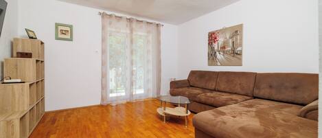 A living room with a brown sofa, wooden flooring, a glass coffee table, a wooden bookshelf, a window with curtains, and a wall-mounted picture.