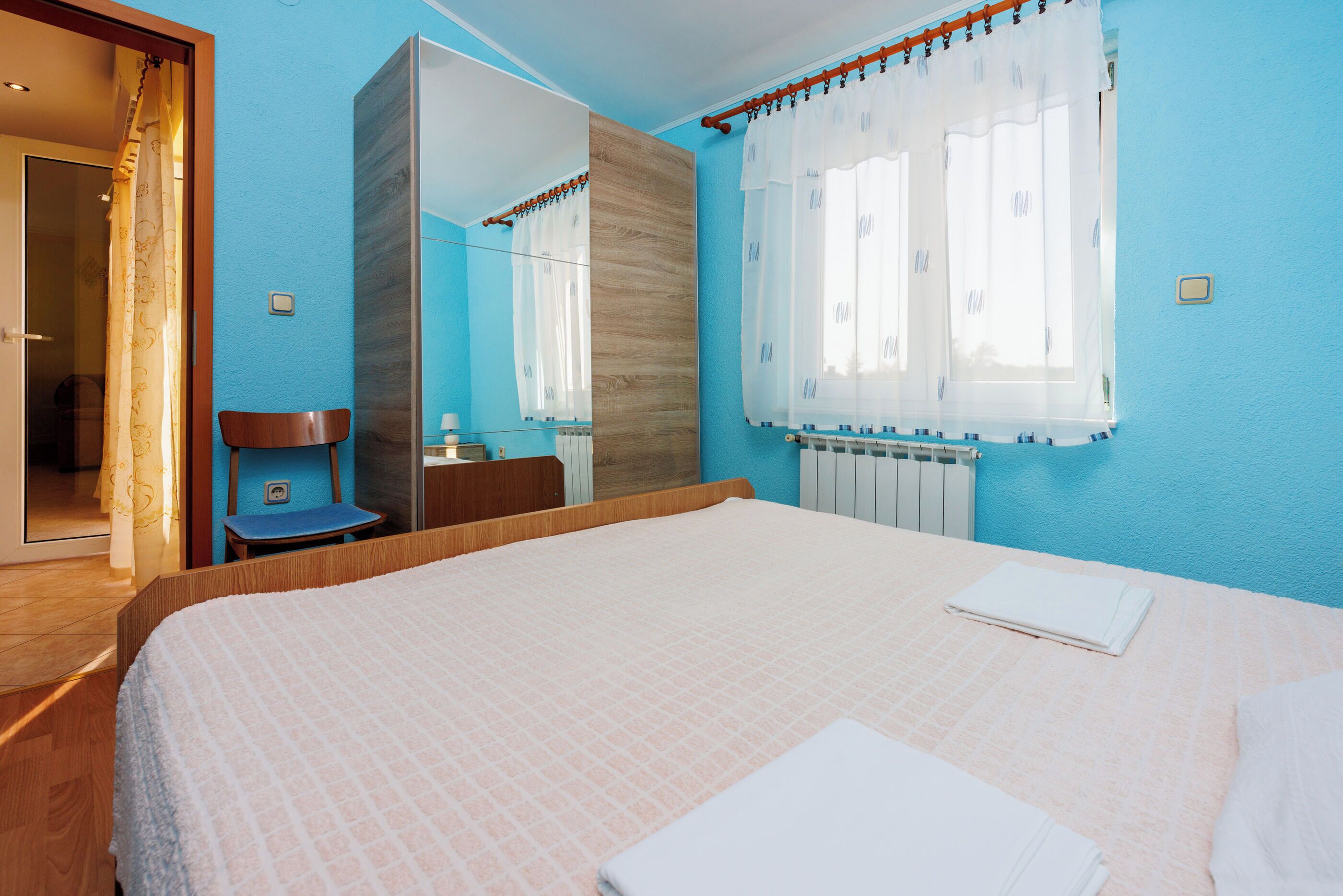 A bedroom with a large bed, a chair, a mirror, and a window with curtains.