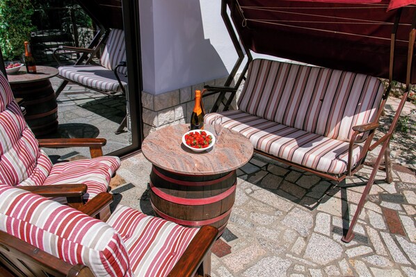 Un espace patio avec des sièges à coussins à rayures, une table ronde avec une bouteille et un bol, et un parasol rouge.