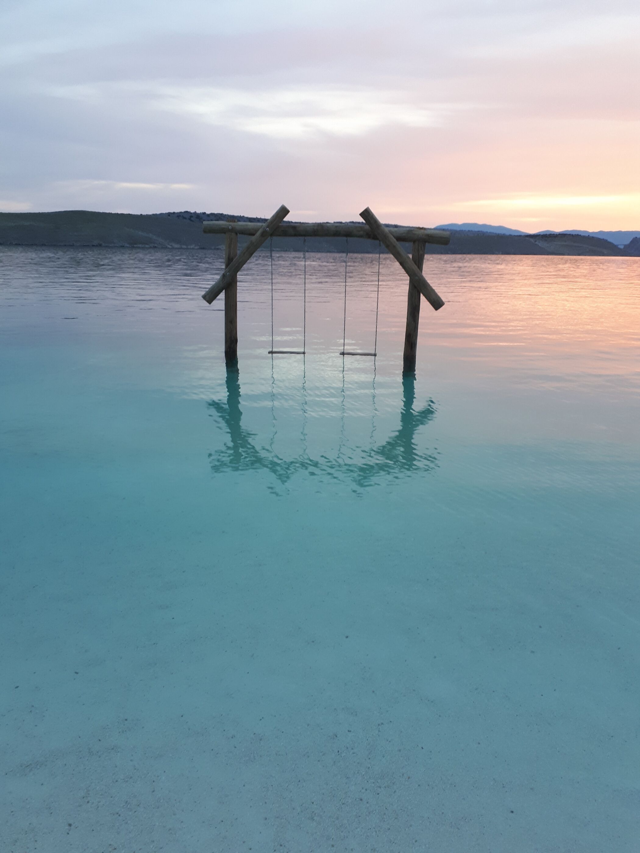 A wooden swing set over a clear blue body of water with a hilly landscape in the background.