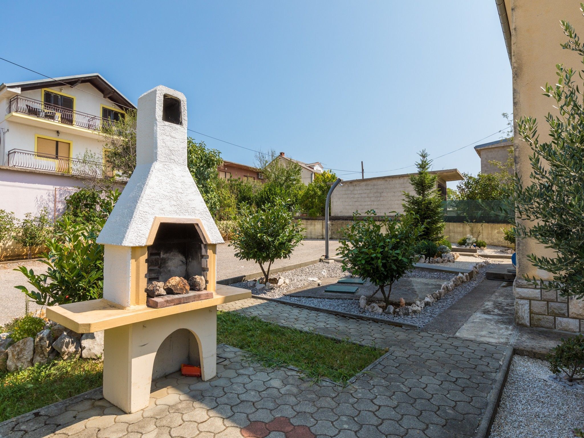 Un forno in pietra esterno con canna fumaria, circondato da un patio lastricato e piccoli alberi.