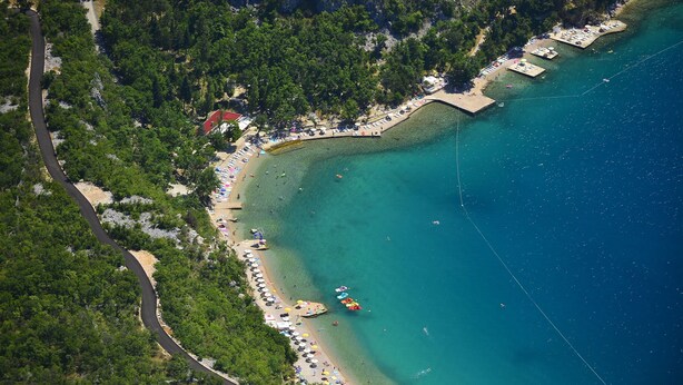 An aerial view of a coastal area with a road curving through a forest, leading to a beach with umbrellas and people. - Apartments Ljiljana (Dramalj)
