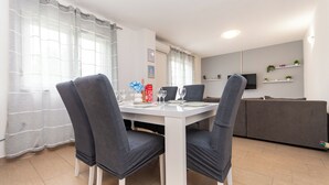 A dining area with a white table and chairs, a TV on the wall, and a grey sofa in the background. - Apartment Cherry Garden (Zemunik Donji)