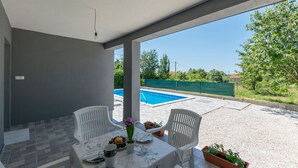 A covered patio area with a table set for two, chairs, and a view of a swimming pool. - Apartment Cherry Garden (Zemunik Donji)