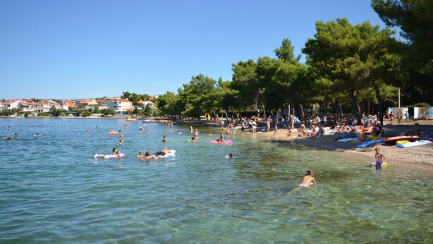 A beach with clear water, people swimming and sunbathing, and a row of houses in the background. - Apartment Cherry Garden (Zemunik Donji)