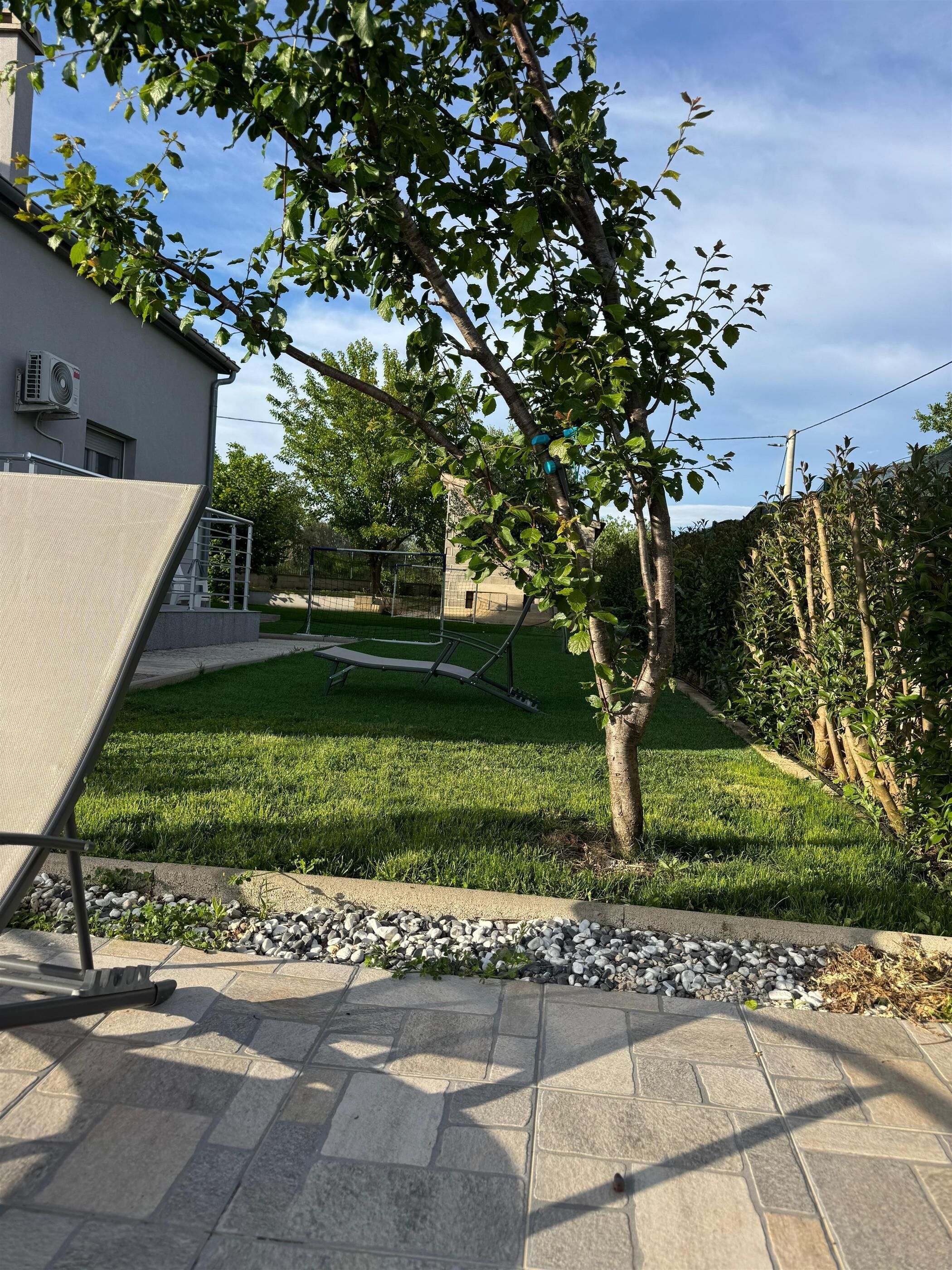 A residential backyard with a lawn, a tree, a metal fence, and a building with an air conditioning unit.