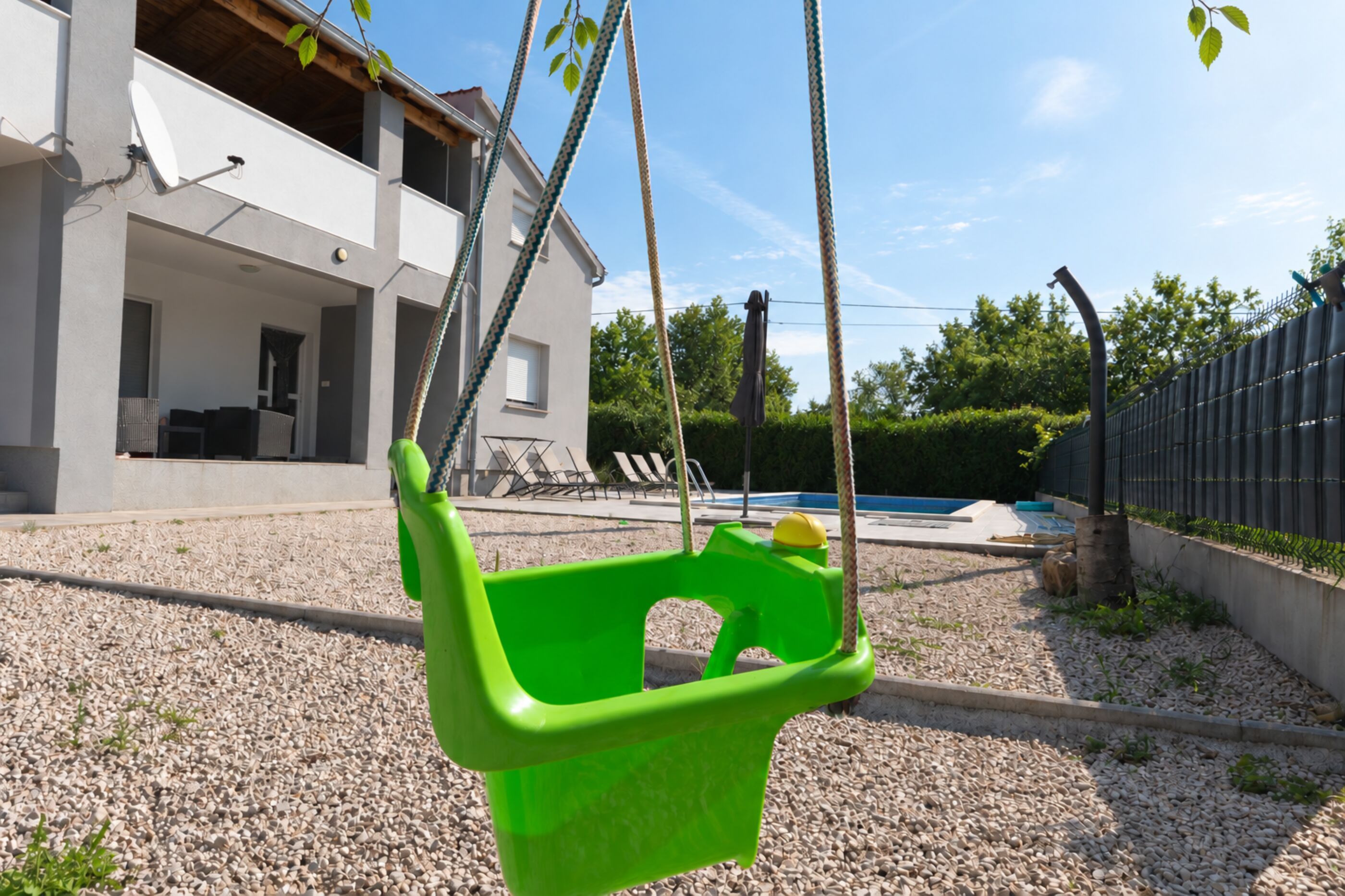 Ein grüner Plastik-Schaukelstuhl in einem Garten mit Swimmingpool, einem weißen Haus und einem Metallzaun.