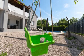 Een groene plastic schommelstoel in een achtertuin met een zwembad, een wit huis en een metalen hek.