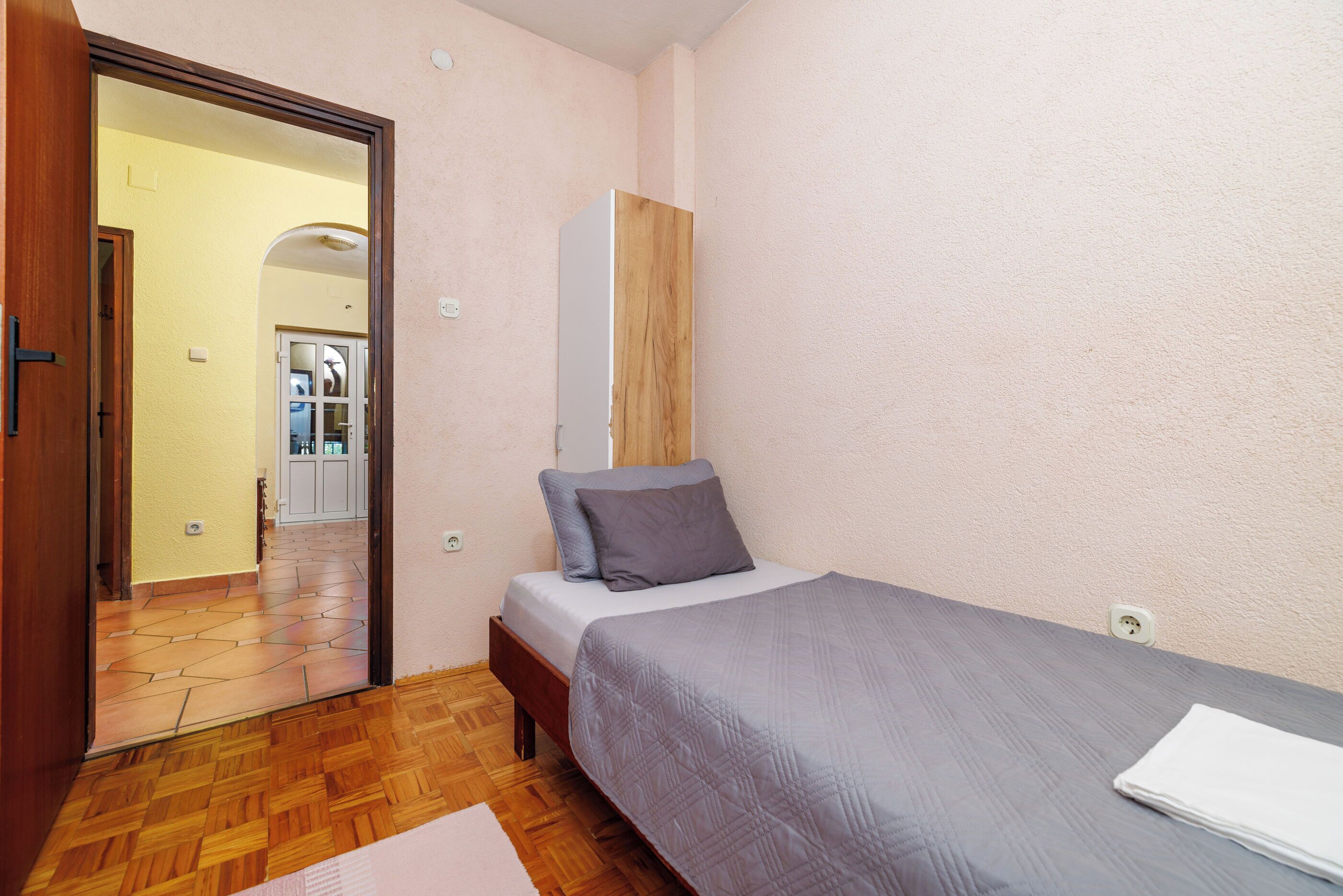 A bedroom with a wooden floor, a bed with a grey bedspread, a wooden wardrobe, and a small white table.