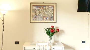 Une pièce avec une table et une chaise blanches, un tableau encadré au mur et un téléviseur fixé au mur.