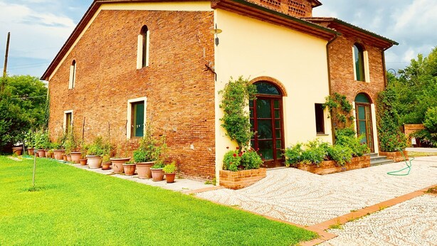 A two-story brick building with a yellow facade, a central arched entrance, and a small porch area with potted plants. - La Fornace del Sole (Capannori)