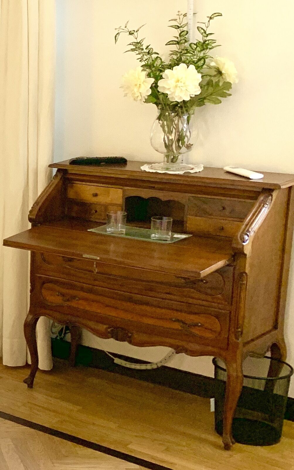 A wooden desk with a glass top, a vase with white flowers, and a green plant on the floor.