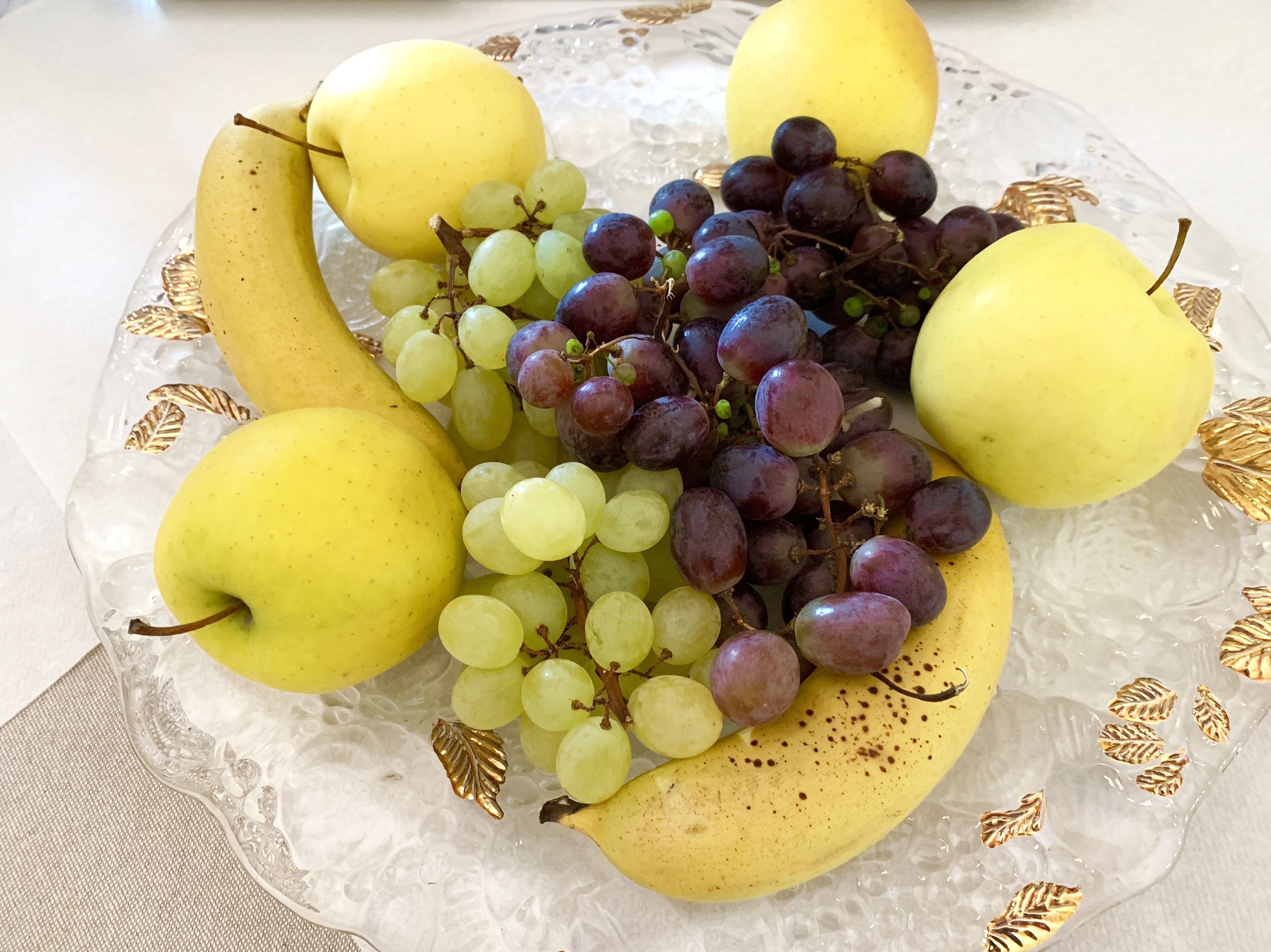 Una bandeja de frutas con plátanos, uvas y peras sobre un plato de vidrio decorativo.
