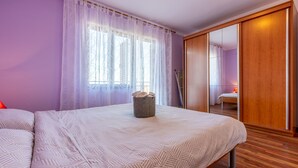 Un dormitorio con una cama, un armario de madera, un ventanal con cortinas y una mesita de noche con lámpara.