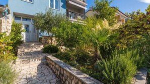 A multi-story building with a stone pathway leading to a door, surrounded by lush greenery and a palm tree. - Apartments Brankica (Dramalj)