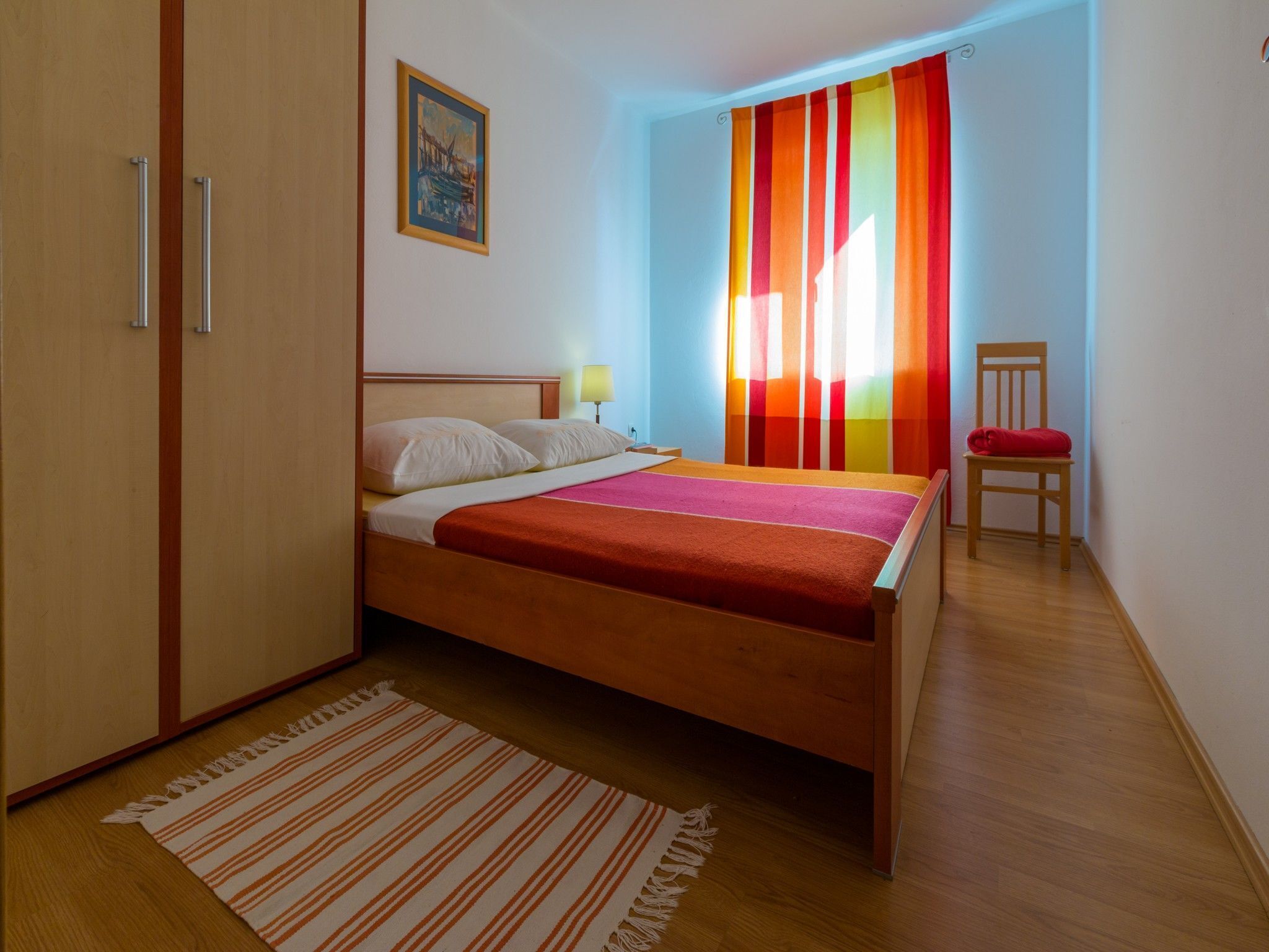 Ein Schlafzimmer mit einem Bett, einem Sessel, einem Fenster mit bunten Vorhängen, einem Bild an der Wand und einem Holzboden.