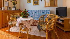 Un salon chaleureux avec un canapé, deux fauteuils en osier, une petite table ronde recouverte d’une nappe en dentelle, un téléviseur et un miroir décoratif.