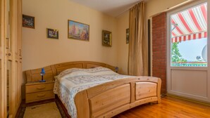 Un lit en bois avec une couvre-lit à motifs, une table de chevet et une petite table d’appoint. On peut voir des tableaux accrochés au mur et une fenêtre surmontée d’un auvent à rayures.